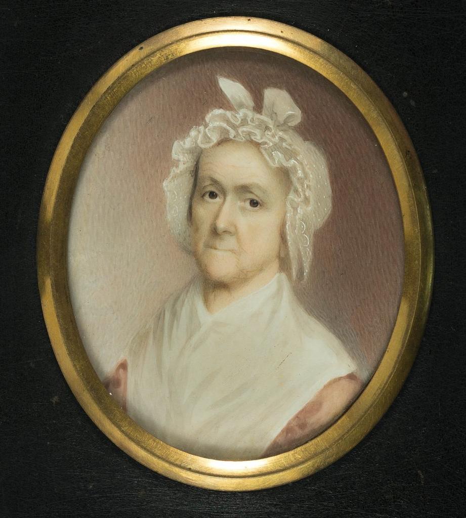 Color photograph of a small portrait painting of an older white woman in an oval gold frame, with black painted wood frame around that. The figure is in ¾ profile and looks at the viewer with expressive dark eyes, she wears a frilled white cap that covers her hair, a large white shawl and a pink dress or shirt. The background is pink, lighter to the right, and gets darker as it goes left.